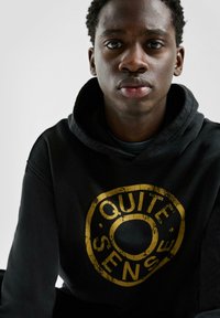 Schwarzer Kapuzenpullover mit einem großen, abgenutzten goldenen, kreisförmigen Logo mit der Aufschrift "QUITE SENSE" auf der Vorderseite, ausgestattet mit einer Kordelzugkapuze.