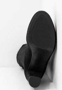 Stivale alla caviglia in suede nero con punta a forma di cono, tacco impilato e motivo a coste sulla suola. I dettagli delle cuciture laterali sono visibili.