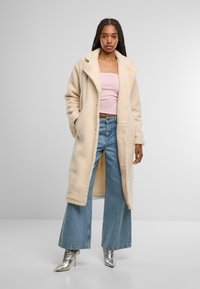 Beige, flauschiger langer Mantel mit Reverskragen, kombiniert mit einem hellrosa Oberteil und weiten blauen Jeans. Silberne High Heels runden das Outfit ab.