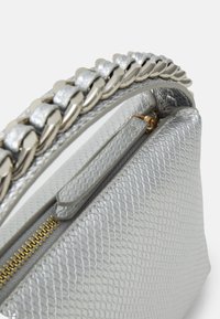 Stříbrná kabelka s texturou, vybavená kovovým řetízkem, zipovým uzavřením a hladkým vnitřním podšívkou. Na vnějším povrchu má výrazný vzor připomínající plaz.