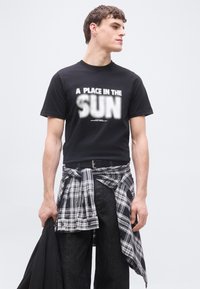 Νέος άνδρας που φοράει μαύρο μπλουζάκι με το κείμενο "Μια θέση στον ήλιο", με καρό πουκάμισο δεμένο γύρω από τη μέση, κρατώντας μαύρο μπουφάν, κοιτώντας προς τα πλάγια.