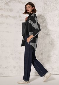 Zwart en grijs geruite cardigan met open voorkant, gedragen over een zwart topje, gecombineerd met navy wijdere jeans en witte sneakers.
