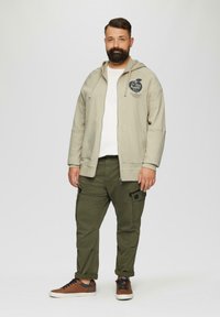 Beige zip-up hoodie met logo, witte t-shirt eronder, olijfgroene cargobroek en bruine sneakers met marineblauwe veters. Casual, relaxte pasvorm.