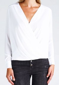 Blusa bianca avvolgente con maniche lunghe, scollatura a V profonda e texture liscia, abbinata a jeans in denim scuro con due bottoni frontali.