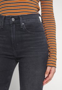 Jeans grigio scuro a vita alta con una texture liscia, design a cinque tasche e chiusura con bottone. Maglietta a maniche lunghe a righe arancione e nera.