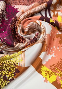 Tela de seda con un patrón en espiral que presenta diseños florales en púrpura, rosa, naranja y amarillo sobre un fondo crema. Textura suave.