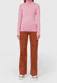 Person, die einen figurbetonten rosa Rollkragenpullover, braune weite Hosen und weiße Sneakers mit grünen und pinken Akzenten trägt.