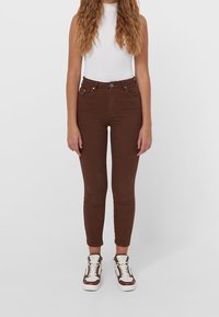 Jeans ajustados marrones de cintura alta, con cinco bolsillos y costuras sutiles, combinados con una camiseta blanca ajustada y zapatillas marrones.