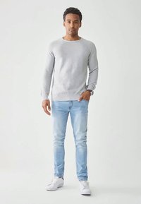 Hellgrauer Strickpullover, blau verwaschene Jeans, weiße Sneakers. Lässiges Outfit mit gerippten Bündchen und Taillenbund, Modell steht vor neutralem Hintergrund.