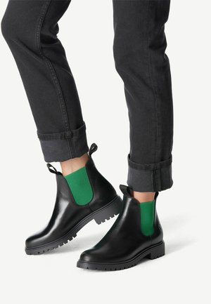 Personne portant des bottines noires avec des panneaux latéraux élastiques verts et un jean noir avec revers, debout devant un fond blanc.