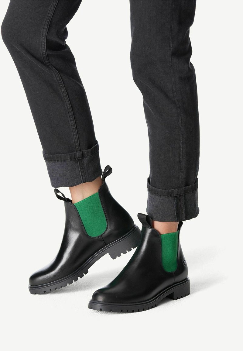 Personne portant des bottines noires avec des panneaux latéraux élastiques verts et un jean noir avec revers, debout devant un fond blanc.