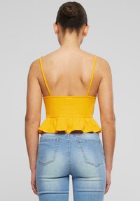 Žlutý peplum top s tenkými ramínky, žebrovanou texturou a rozšířeným lemem, kombinovaný se světle modrými džínami s vysokým pasem, viděný ze zadní strany.