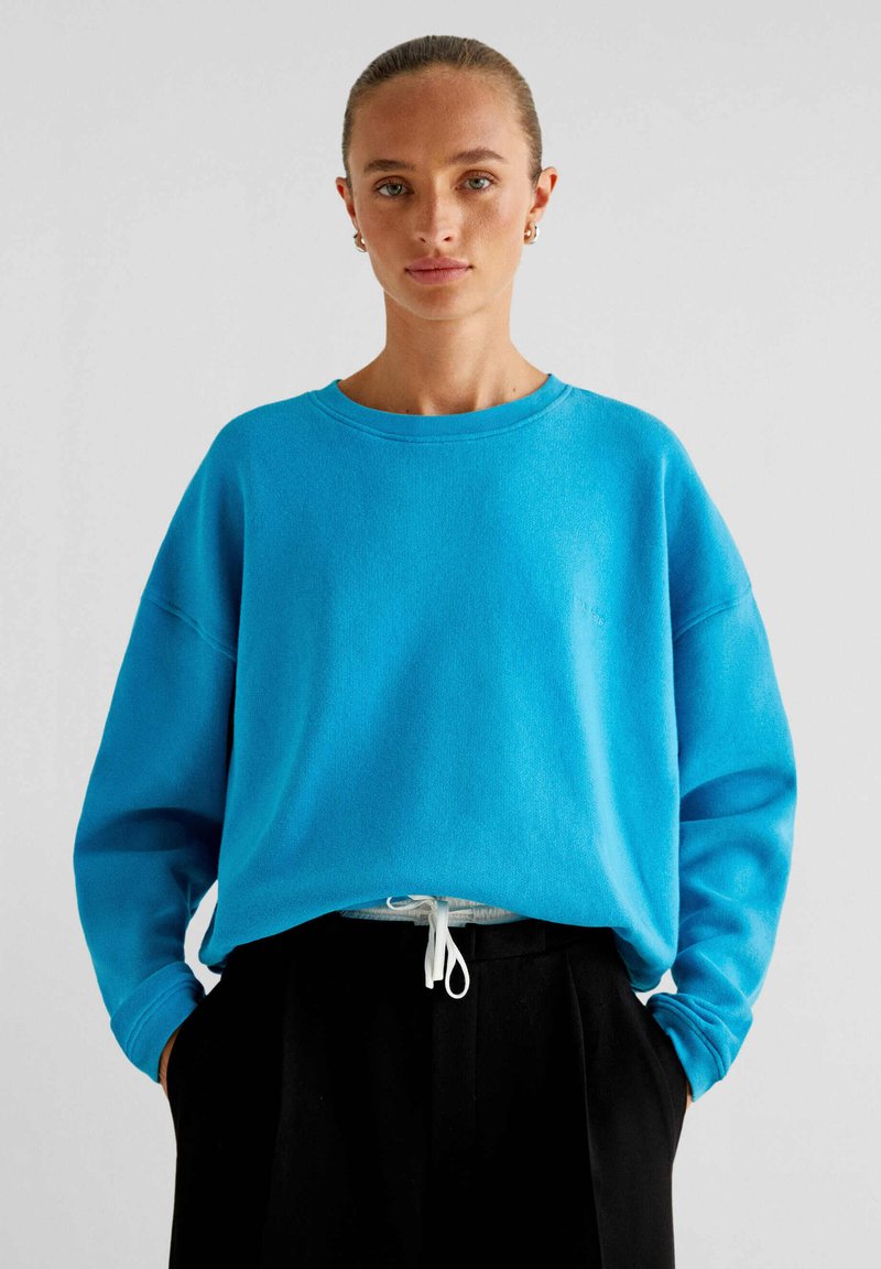 Blauwe sweatshirt met een ronde hals en een oversized ontwerp, met lange mouwen en een witte koord bij de taille, gecombineerd met zwarte broek.