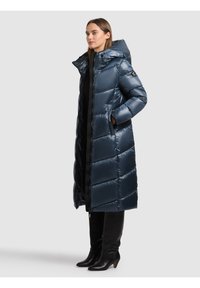 Kvinde iført en lang, skinnende navyblå quiltet dynejakke med høj krave og hætte, sorte bukser og sorte støvler med hæl.