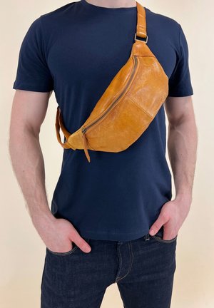 Homme portant un t-shirt bleu marine et un jean foncé avec un sac en cuir marron clair porté en bandoulière, les mains dans les poches.