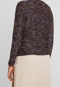 Giacca lavorata a maglia in tonalità scure con accenti blu e rossi. Dotata di maniche lunghe e taglio dritto, adatta per essere indossata a strati sopra gli outfit.