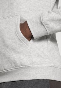 Grijze hoodie met een kangoeroezak, ribbelboorden en textuurstof. De close-up benadrukt de naaideetails en materiaaleigenschappen.