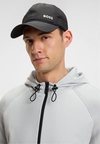 Egy férfi fekete sapkát visel, amelyen a "BOSS" felirat található, valamint egy világosszürke zipzáros kapucnis pulóvert, amelynek zsinórjai vannak. A férfi enyhén balra fordulva áll egy fehér háttér előtt.