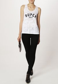 Witte mouwloze tanktop met zwarte "SUPER DRY" print, gecombineerd met zwarte skinny jeans en zwarte schoenen, en een grijze clutch in de hand.