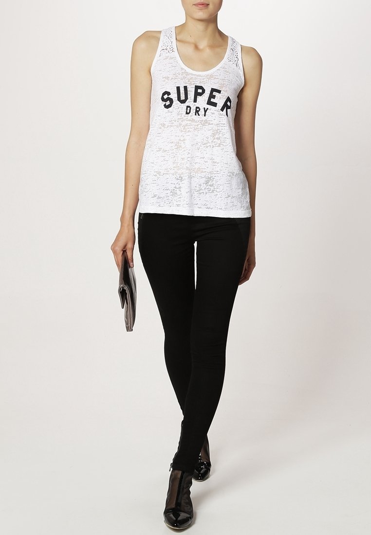 Witte mouwloze tanktop met zwarte "SUPER DRY" print, gecombineerd met zwarte skinny jeans en zwarte schoenen, en een grijze clutch in de hand.