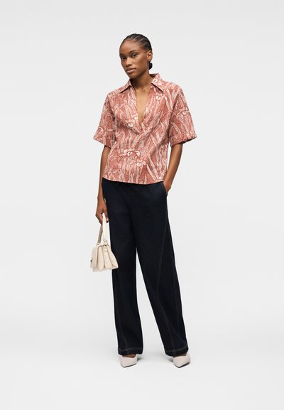 Femme aux cheveux tressés portant un chemisier à manches courtes à motifs rouille, un pantalon noir à jambes larges, des talons blancs et tenant un sac à main blanc.