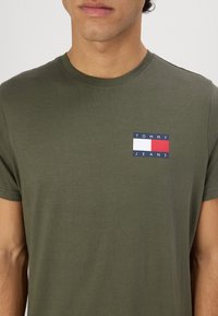 Bavlněné tričko olivově zelené barvy s kulatým výstřihem. Na přední straně je obdélníkové logo v červené, bílé a modré barvě s nápisem "TOMMY JEANS".