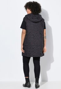 Chaleco acolchado negro con capucha, que presenta un patrón floral texturizado en rojo oscuro. Combinado con una camiseta negra de manga corta y jeans ajustados.