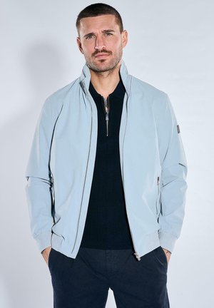 Mann mit kurzem Haar und Bart, der eine hellgraue Reißverschlussjacke über einem schwarzen Pullover trägt, steht mit den Händen in den Taschen vor einem schlichten Hintergrund.