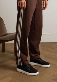 Pantaloni sportivi marroni con strisce bianche sui lati abbinati a scarpe nere con suola bianca e occhielli metallici. Sedia di legno sullo sfondo.