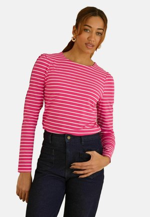 Žena s dlhými vlasmi, v náušniciach kruhoch, oblečená v ružovo-bielom pruhovanom tričku s dlhými rukávmi a tmavých džínsoch s vysokým pásom, na bielom pozadí.