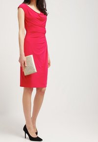 Tajt, knälång klänning i ljusrosa tyg med draperad halsringning. Matchas med en metallisk guldclutch och svarta högklackade skor.