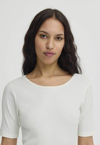 Jeune femme aux longs cheveux ondulés et foncés, portant un haut blanc à manches courtes côtelé, faisant face à l'avant avec une expression neutre.