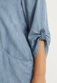 Hellblaues Denim-Hemd mit aufgerollten Ärmeln, einem Knopfdetail und abgerundetem Saum. Der Stoff hat eine weiche, strukturierte Optik.