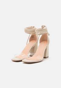 Beige högklackade sandaler med en klar rem, öppen tå och tjock klack. Har korskorsade ankeln band och en slät textur.