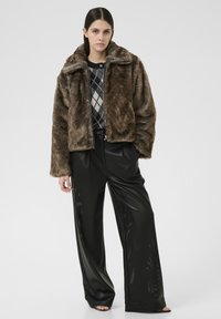 Veste en fausse fourrure marron avec un large col, associée à un pantalon large noir brillant. L'ensemble présente un pull à carreaux en dessous.