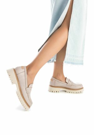 Mocasines de cuero beige claro con una suela blanca gruesa. Presentan un diseño de costura y una correa decorativa en la parte superior.