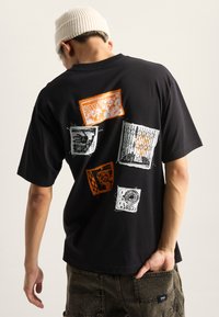 Sort bomulds t-shirt med flerfarvede grafiske tryk på ryggen, med orange, hvidt og gråt design i forskellige former og mønstre.