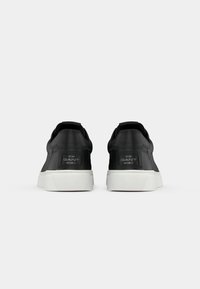 Sneakers negre din piele cu tălpi de cauciuc alb, având un design low-top, o bandă de tragere în spate și un etichetă de marcă embosată pe călcâi.