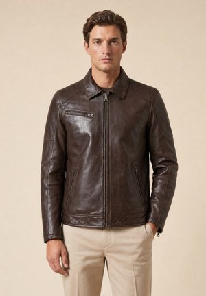 Mann trägt eine dunkelbraune Lederjacke mit Reißverschlusstaschen und beigefarbene Hose, steht vor einem schlichten beigen Hintergrund.