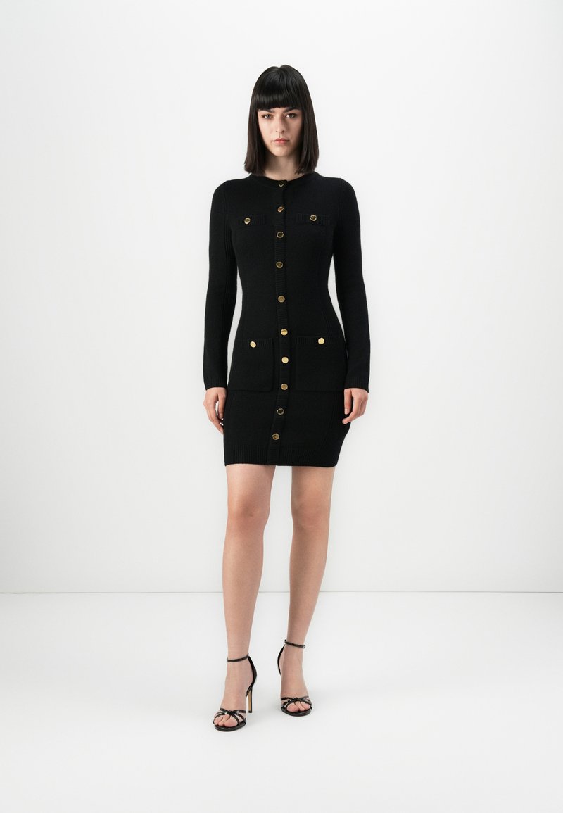 Donna in un abito in maglia nero con bottoni, maniche lunghe e quattro tasche frontali, indossa sandali con tacco alto neri, su sfondo bianco.