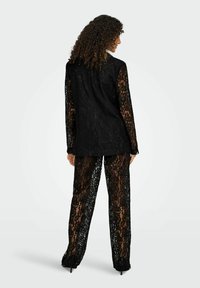 Črno čipkasto obleko sestavljata jakna z dolgimi rokavi in usklajene hlače. Zapleten cvetlični vzorec, prosojna tekstura, krojena prileganje, elegantna silhueta.