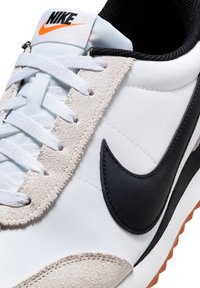 Sneaker Nike con tomaia in tessuto bianco, swoosh in pelle nera, dettagli in suede beige e lacci bianchi con un logo arancione sulla linguetta.