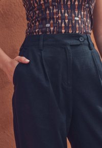 Mørkeblå højttaljede bukser med plisseringer, detaljeret taljebånd og sidelommer. Stoffet har en glat, let struktureret overflade.