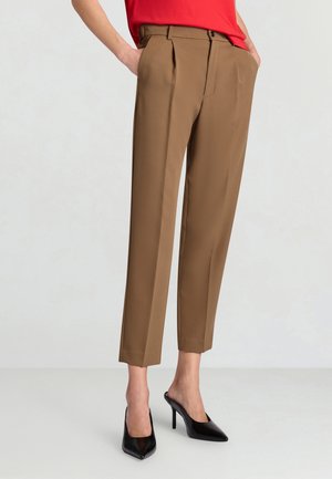 Braune maßgeschneiderte, knöchellange Hose mit vorderen Falten, getragen mit schwarzen spitzen, hochhackigen Mules und einem roten Oberteil.