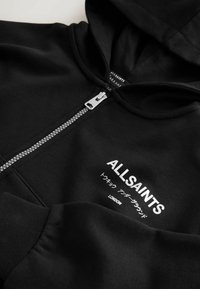 Crna zip-jakna izrađena od mekog materijala. Ima prednji patentni zatvarač, kengurovu džep i bijeli tekst s japanskim karakterima na prsima.