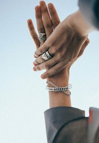 Silverringar i olika mönster på händer, inklusive staplade band och geometriska former. Ett silverkedje-armband pryder en handled.