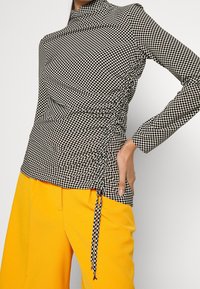 Černobílý kostkovaný top s dlouhým rukávem a nařasenými boky, s vysokým límcem a detaily s šňůrkou, spárovaný s jasně žlutými kalhotami.