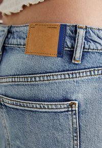 Light blue denim jeans with orange stitching, featuring a leather patch near the waistband. The fabric shows a soft, textured weave.