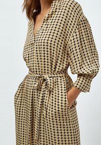 Beige mekko, jossa on musta geometrinen kuvio, lyhyet hihat ja vyötäröllä vyö. Kangas on sileä ja siinä on sivutaskut.