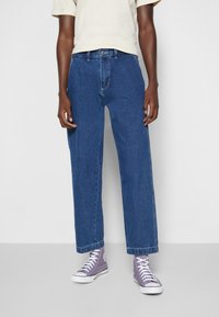 Blue denim jeans with a relaxed fit, featuring a button and zipper closure, side pockets, and contrasting stitching; worn with purple high-top sneakers.